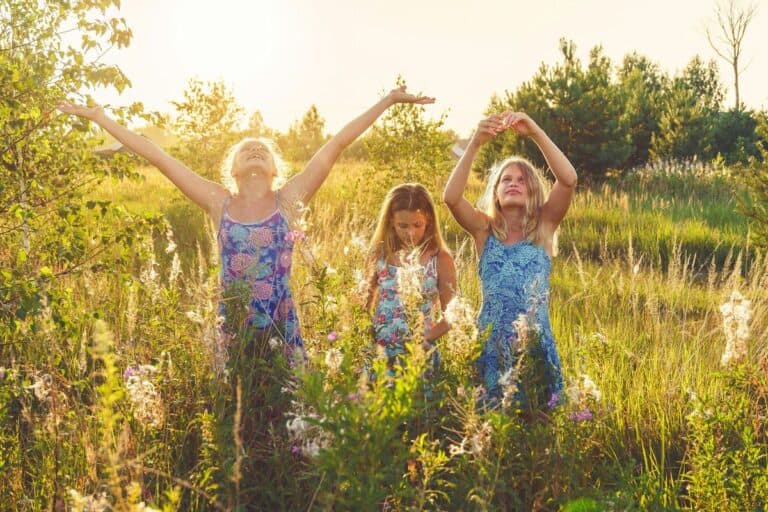 Two girls and a young woman joyfully stretching their arms towards the sun in a lush meadow, embodying the spirit of environmentally aware children enjoying nature's beauty.