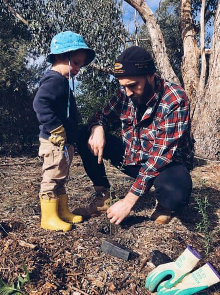 A young child in a blue hat and yellow boots attentively watches an adult in a plaid shirt and beanie plant a seedling, with gardening tools laid aside on mulched soil, engaging in an educational moment about environmental stewardship.