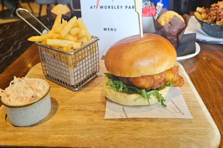 A delicious meal served at Tempo at Worsley Park. The dish features a crispy fish sandwich with fresh rocket on a soft bun, accompanied by a side of golden fries in a metal basket and a small bowl of creamy coleslaw. A menu card with the restaurant's name is visible in the background.