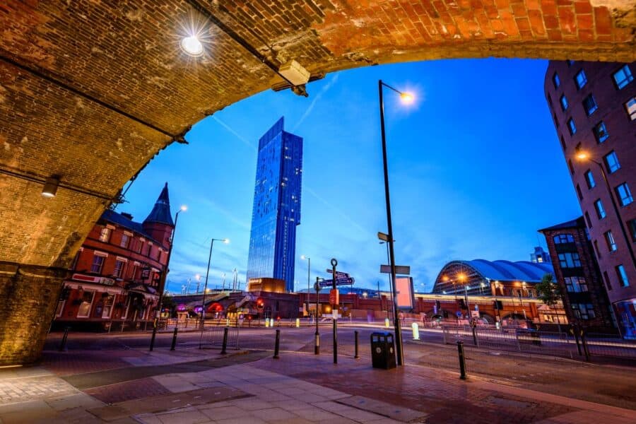 Night view of Manchester's cityscape, framed by a brick archway, with the Beetham Tower and Manchester Central visible under a deep blue sky.