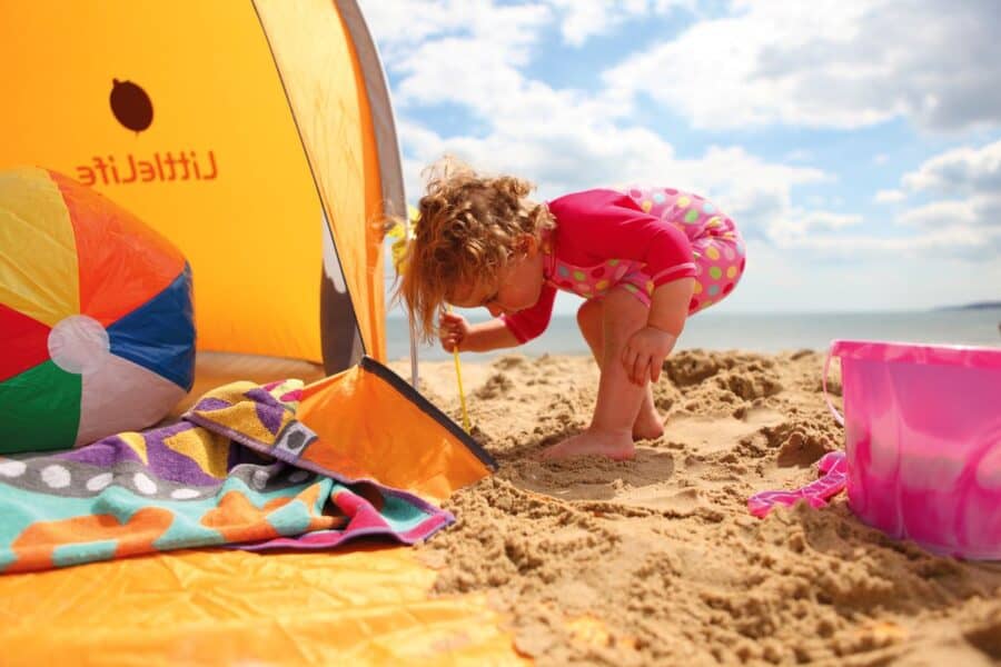 Little girl playing with beach toys in front of a grey and orange beach tent with a curved roof and open front entrance.
