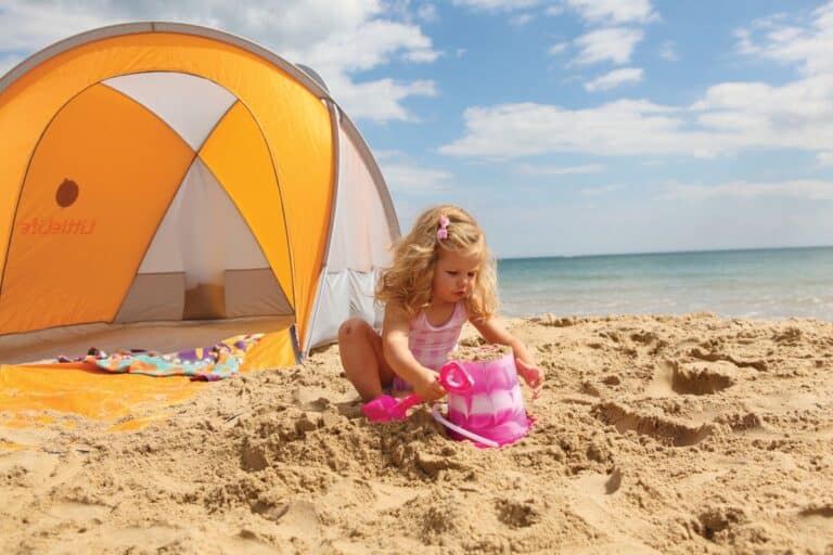 LittleLife Family Beach Shelter review - A child playing with a sand bucket in front of a grey and orange beach tent set up on a sandy beach, demonstrating the shelter’s spacious and family-friendly design.