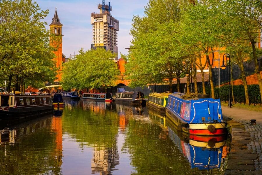 Colourful narrowboats moored along the River Irwell in Manchester, with lush green trees and historic buildings reflected in the calm water under a clear sky.