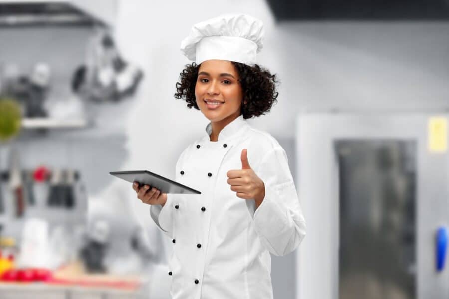 Smiling executive chef wearing a professional white double-breasted jacket and hat, holding a tablet in a modern kitchen.