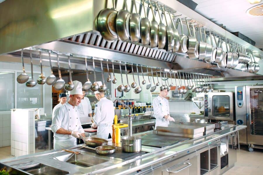 Professional chefs wearing white uniforms and hats, working in a commercial kitchen with stainless steel appliances and hanging utensils.