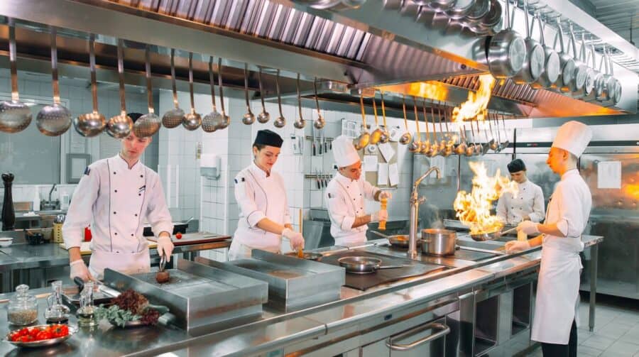 Professional chefs in a commercial kitchen, wearing white uniforms and hats, cooking with flames and preparing ingredients in a fast-paced environment.