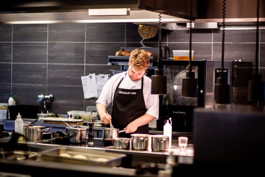 Professional chef wearing a black apron over a white chef jacket, whisking ingredients in a saucepan in a modern kitchen.